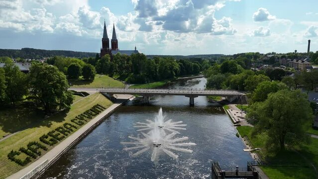 Aerial view of Anyk&scaron;čiai, Lithuania, featuring the church, a decorative fountain in the river, and lush greenery under a vibrant blue sky.