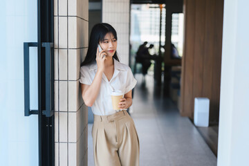 Young Woman Talking on Phone While Holding Coffee Cup in Modern Office Building with Natural Light