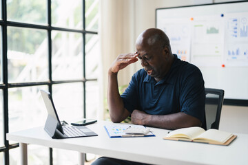 Senior Businessman Working at Desk with Laptop and Documents in Modern Office, Analyzing Financial Data, Stressful Work Environment, Daylight through Large Windows