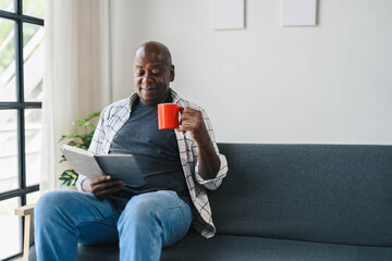 Senior Man Enjoying a Relaxing Moment at Home with a Book and Coffee Mug, Sitting on a Comfortable Sofa in a Bright, Modern Living Room with Natural Light and Minimalist Decor