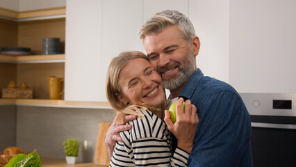 Adults happy family 40s smiling positive couple middle-aged man woman at kitchen cuddling hug embracing food delivery order wife give apple husband embrace hugging at kitchen cuddle love bonding