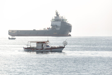 Small traditional Cypriot fishing boat sailing near a big cargo ship in the sea at Akrotiri Bay, Cyprus