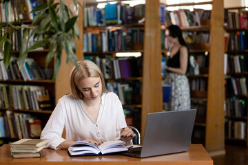 a girl sits at a table in a library, reading a book and taking notes. There is also a laptop and several closed books on the table.