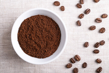 Coffee powder, freshly ground coffee beans in a white bowl on linen fabric. On the right roasted seeds of berries from Coffea arabica, also Arabian, mountain or arabica coffee. Close-up, from above.
