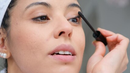 A woman is applying mascara to her eyes. She is wearing a white towel around her head