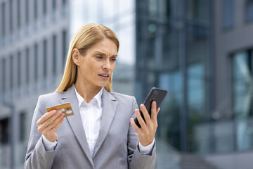 Fototapeta premium Businesswoman with a worried expression holding a credit card and smartphone. She is standing outside an office building, suspecting possible fraud or financial issue.