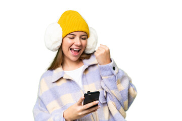 Young blonde woman wearing winter muffs over isolated chroma key background with phone in victory position