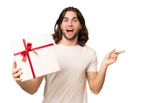 Young handsome man holding a gift over isolated background surprised and pointing finger to the side