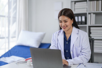 Female doctor using a laptop in a medical consultation room, wearing a white coat and professional attire, smiling while working.