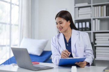 Female doctor working on a laptop while taking notes on a clipboard in a modern medical office. Healthcare professional in a clinical setting.