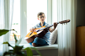 Kid playing guitar. Child boy sitting on the windowsill and playing acoustic guitar