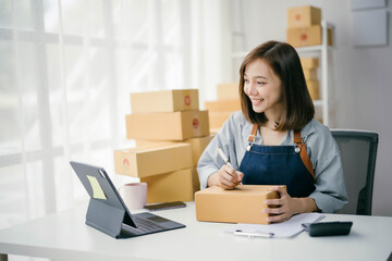 Young Woman Working from Home, Managing Online Business Orders, Writing on Packages, Using Tablet, Surrounded by Cardboard Boxes, Bright Workspace, E-commerce Concept, Small Business Owner