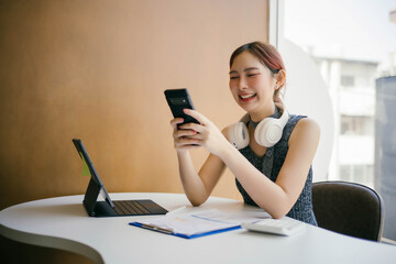 Young Woman Smiling While Using Smartphone at Modern Workspace with Tablet and Headphones, Casual Business Setting, Bright and Cheerful Atmosphere, Productive Work Environment