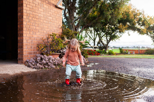 happy toddler with pigtails jumping in puddle of rain water on farm