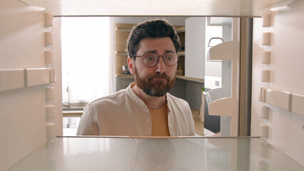 Hungry Caucasian bachelor man looking for food in empty fridge finding no meal products feeling sad...