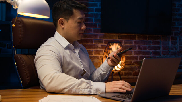 Asian middle-aged male office worker at night evening Korean man businessman executive with laptop working dictate business audio message voicemail on phone answer with vocal AI smartphone technology