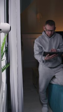 Caucasian man sets up security camera at home for the night. He turns on night vision on CCTV camera using digital tablet and application. Concept of monitoring system, safety and personal privacy.