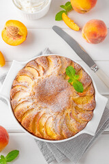Delicious peach pie in a white ceramic baking form on a white wooden background. Summer cake. Selective focus, copy space.