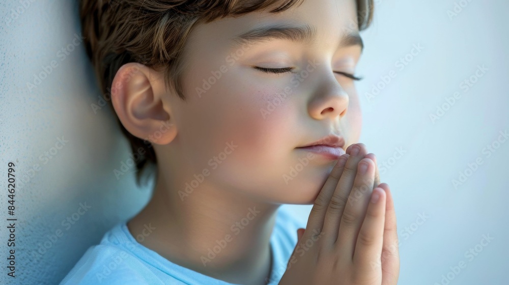 Wall mural a young boy with closed eyes hands clasped together in prayer standing against a light blue backgrou