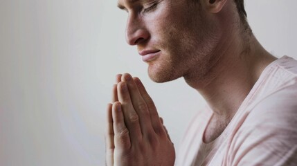 Fototapeta premium A man with a beard and closed eyes clasping his hands together in a gesture of prayer or meditation wearing a light-colored shirt against a neutral background.