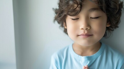 Young child with eyes closed possibly in a state of relaxation or prayer wearing a light blue shirt with soft curly hair against a neutral background.