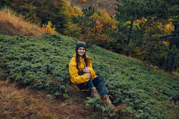 Young woman in stylish yellow raincoat sitting on grassy hillside in beautiful autumn scenery, enjoying nature and travel