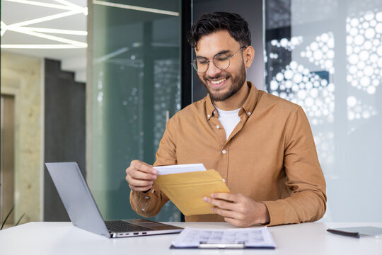Happy indian man sitting at desk in modern office and smilingly opening and reading received letter