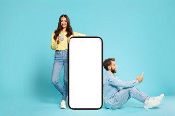 Young European couple near oversized smartphone blank screen, ideal for showcasing apps or websites, man and woman using their cellphones