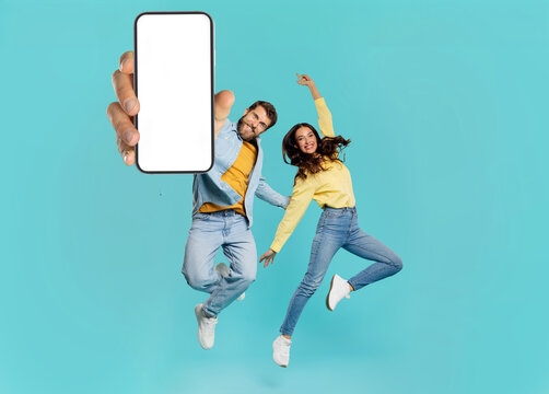 Great news. Overjoyed European couple showing huge smartphone with white blank screen, jumping and celebrating over blue. Mockup collage