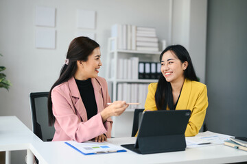 Two Professional Women Collaborating in Modern Office Setting with Laptop and Documents, Engaging in Productive Discussion and Smiling, Business Meeting, Teamwork, Corporate Environment