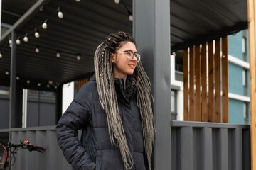 A pretty young woman with a dreadlocked hairstyle dressed in a winter jacket walks around the city