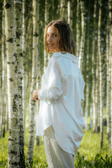 A woman with short hair smiles warmly while standing in a birch tree forest. She is wearing a white shirt, and the sunlight filters through the trees, creating a cheerful and natural atmosphere.