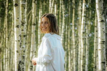 A woman with short hair smiles warmly while standing in a birch tree forest. She is wearing a white shirt, and the sunlight filters through the trees, creating a cheerful and natural atmosphere.