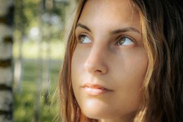 Close-up of a womans face with green eyes, showing a natural and serene expression. The background features blurred greenery, enhancing the focus on her facial features.