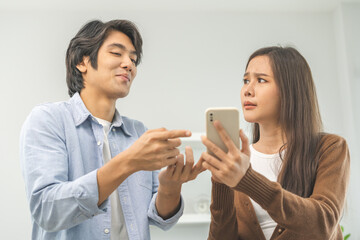 Fototapeta premium Infidelity, suspicion asian young couple love fight relationship, wife holding cellphone, smartphone cheating on phone, scolding husband about mistrust, distrust and jealousy at home.