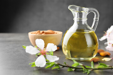 Almond oil in jug, branch with flowers and nuts on grey textured table, closeup. Space for text