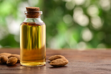 Almond oil in bottle and nuts on wooden table against blurred green background, closeup. Space for...