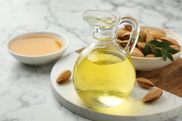 Almond oil in jug, nuts and leaves on white marble table, closeup