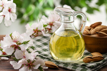 Almond oil in jug, flowers and nuts on wooden table against blurred green background, closeup