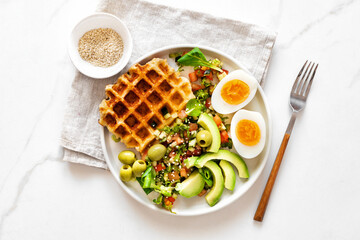 Healthy breakfast or brunch featuring boiled eggs, salad with avocado, olives and cheese waffles on white marble background top view