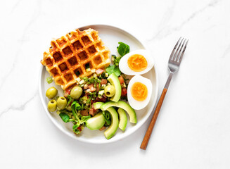 Top view healthy breakfast or brunch featuring boiled eggs, salad with avocado, olives and cheese waffles on white marble background