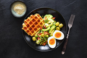 Top view healthy breakfast or brunch featuring boiled eggs, salad with avocado, olives and cheese waffles on dark stone background