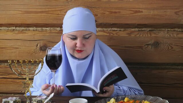 A Jewish woman in a blue veil with glasses of wine and siddur reads kiddush for wine on the Pesach Seder holiday. Medium plan