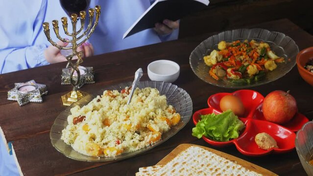 A Jewish woman in a blue veil with glasses of wine and siddur reads kiddush for wine on the Pesach Seder holiday at a set table. Medium plan