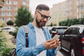 Outdoor portrait of attractive smiling casually dressed bearded man in stylish eyeglasses browsing internet on his smartphone while walking