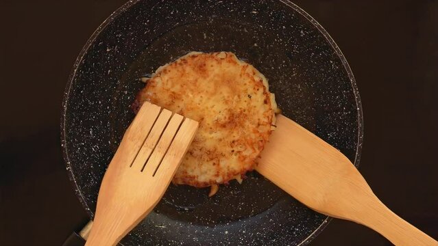 A wooden spatula turns a ruddy fried appetizing potato pancake in a frying pan. High quality 4k footage