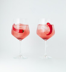 Two wine glasses filled with rose-colored cocktails, each garnished with a chili pepper and ice cubes, set against a white background. The combination of spicy chili and refreshing drink