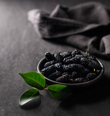 Fresh black mulberry with leaf in black wooden bowl on dark background with napkin. Concept of juicy and healthy seasonal garden berries. Copy space.