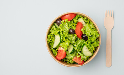 Fresh green salad with tomatoes and cucumbers in a paper container with a wooden fork on a light background. Healthy food concept in biodegradable eco-friendly takeaway dishes. Top view and copy space