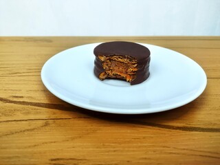 Bitten chocolate alfajor with dulce de leche and coffee on the wooden table
